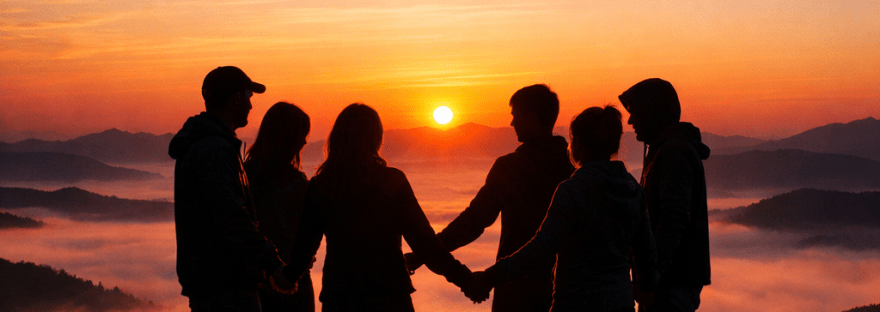 Six people holding hands in a circle on a mountain during sunrise with misty hills in the background