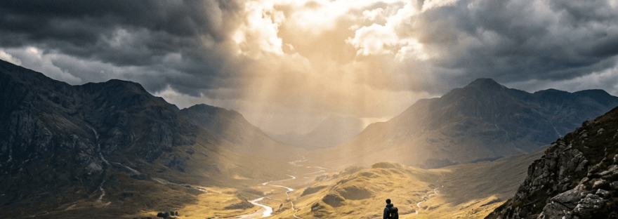 Hiker standing on rocky ridge overlooking valley illuminated by sunrays through clouds