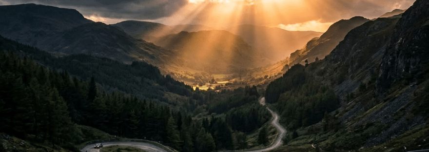 Winding mountain road with cars and sun rays breaking through dark clouds